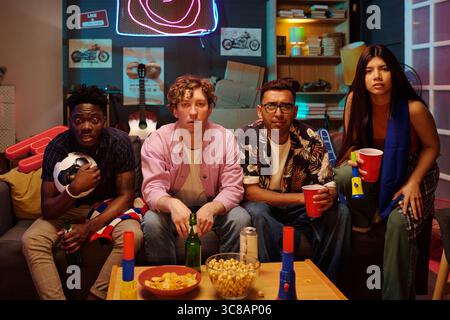 Gruppe multiethnischer junger Erwachsener, die auf der Couch sitzen und intensiv fernschauen, Getränke und Snacks, Fußball- und Schaumhände in der Hand halten und fokussiert auf dem Bildschirm erscheinen Stockfoto