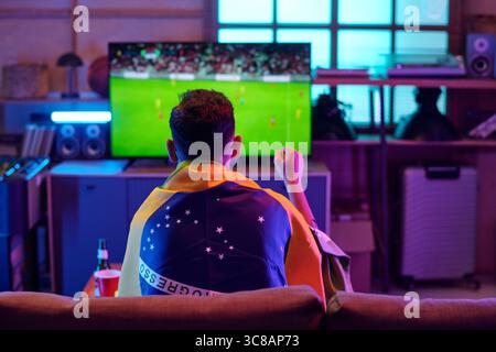 Kaukasischer junger Erwachsener, der auf dem Sofa sitzt und Fußballspiele im Fernsehen ansieht, brasilianische Flagge über den Schultern trägt, vor Aufregung die Faust hält, Getränke auf dem Tisch in der Nähe Stockfoto