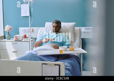 Birassischer Mann mittleren Alters isst Frühstück, während er im Krankenhausbett ruht Stockfoto