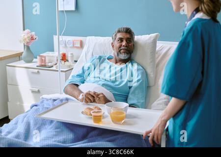 Birassischer Mann mittleren Alters, der im Krankenhausbett liegt und das Frühstück von der Krankenschwester erhält Stockfoto