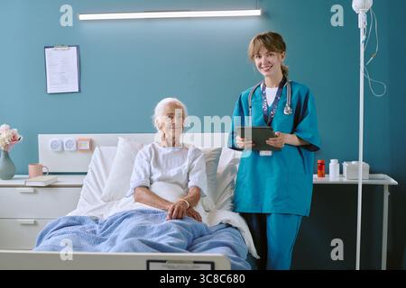 Ältere Frau liegend im Krankenhausbett mit junger Erwachsenenschwester lächelnd Stockfoto