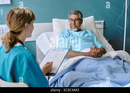 Birassischer Mann mittleren Alters, der im Krankenhausbett liegt und mit weiblicher Ärztin spricht Stockfoto