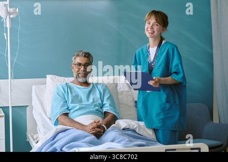 Birassischer Mann mittleren Alters, der im Krankenhausbett sitzt und die junge Erwachsene Krankenschwester lächelt Stockfoto