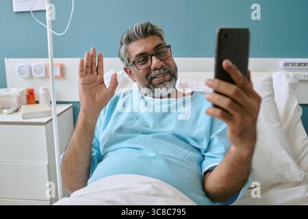 Birassischer Mann mittleren Alters, der im Krankenhausbett liegt und während eines Videogesprächs winkt Stockfoto