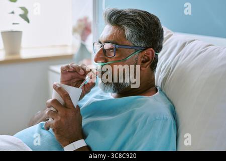 Birassischer Mann mittleren Alters, der im Krankenhausbett ruht und durch Strohhalm trinkt Stockfoto