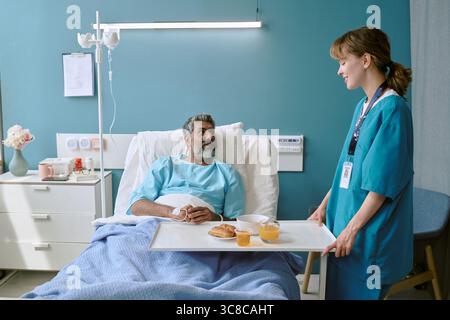 Birassischer Mann mittleren Alters, der im Krankenhausbett liegt und eine Mahlzeit von der Krankenschwester erhält Stockfoto