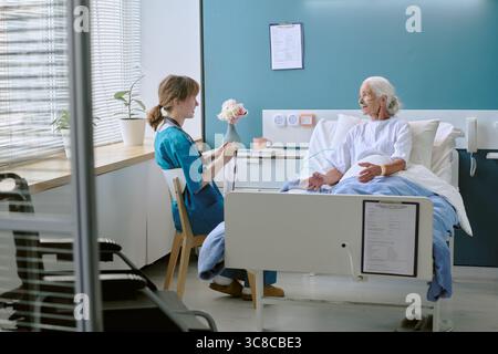 Ältere kaukasische Frau lächelt, während sie mit der jungen Erwachsenen-Krankenschwester spricht Stockfoto