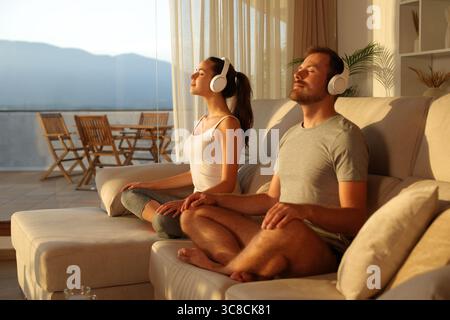 Paare zu Hause machen Yoga mit Kopfhörern, die zu Hause bei Sonnenuntergang auf einer Couch sitzen Stockfoto