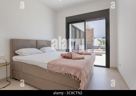Minimalistisches Schlafzimmer mit einem großen beigefarbenen Bett mit frischer Bettwäsche und Handtüchern, neben Schiebetüren, die auf eine private Terrasse mit Sitzgelegenheiten im Freien führen Stockfoto