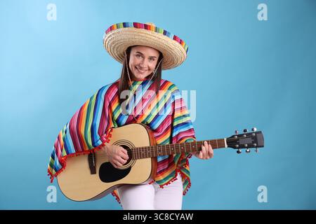 Lächelnde Frau in Sombrero-Hut und farbenfrohem Poncho, der Gitarre auf hellblauem Hintergrund spielt Stockfoto