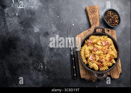 Fusilli Carbonara Nudeln mit knusprigen Speckstückchen und Parmesankäse in einer Pfanne. Schwarzer Hintergrund. Draufsicht. Stockfoto
