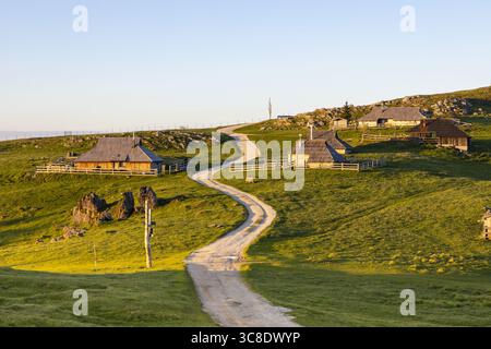 Blick auf eine gewundene Straße schlängelt sich durch grüne Hügel mit traditionellen Holzhütten unter klarem Himmel, Velika Planina, Stahovica, Kamnik, Slowenien. Stockfoto