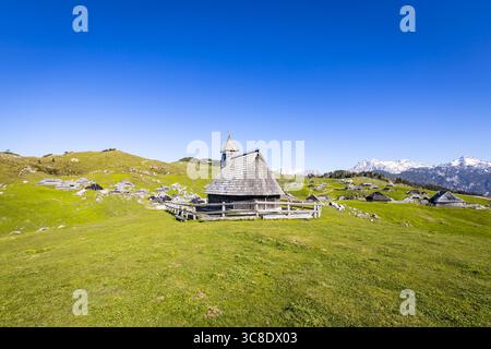 Blick auf eine alte Holzkirche, umrahmt von einem rustikalen Zaun, liegt auf einem lebhaften grünen Hügel vor dem Hintergrund schneebedeckter Berge, Stahovica, Kamni Stockfoto