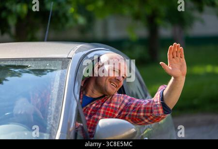 Fröhlicher Mann in kariertem Hemd winkt vom Fahrersitz des geparkten Autos, umgeben von Grün und hellem Sonnenlicht. Stockfoto