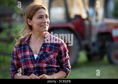 Hübsche Frau in kariertem Hemd lächelt, als sie vor dem Traktor auf dem Bauernhof steht und das ländliche Leben und die landwirtschaftliche Arbeit zeigt Stockfoto
