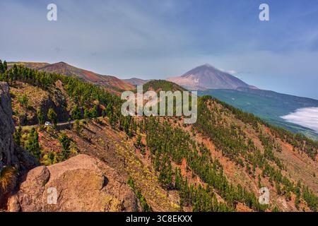 Vulkanische Landschaft mit Pico del Teide im Hintergrund, gewundene Straße und Kiefernwald über felsige Berghänge Stockfoto