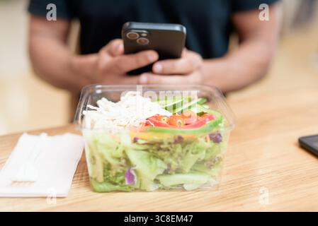 Nahaufnahme einer Hand, die ein Smartphone hält, während sie mit frischem Salat in einem Behälter zum Mitnehmen interagiert. Im Lebensmittelbereich des Einkaufszentrums erschossen. Gesunde Ernährung, mobil Stockfoto