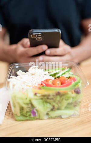 Nahaufnahme einer Hand, die ein Smartphone hält, während sie mit frischem Salat in einem Behälter zum Mitnehmen interagiert. Im Lebensmittelbereich des Einkaufszentrums erschossen. Gesunde Ernährung, mobil Stockfoto