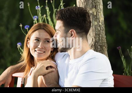 Das junge Paar entspannt sich auf der roten Bank im üppigen Garten, lächelt und teilt am Nachmittag einen Moment der Freude unter der warmen Sonne Stockfoto
