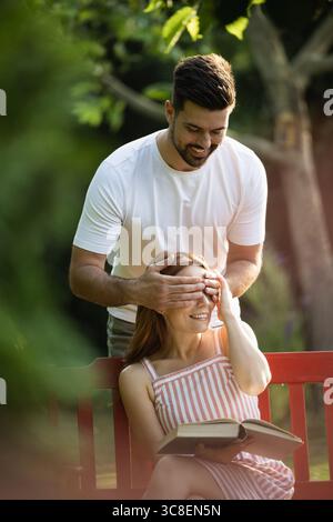 Ein überraschender Mann, der die Augen seines anderen an einem sonnigen Tag im Park bedeckt. Die glückliche Frau las ein Buch, während sie auf einer Bank saß Stockfoto