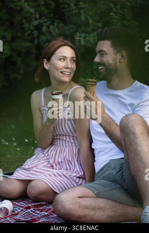 Hübsche Freundin, die ihren Freund liebevoll ansieht, während sie beide Eis essen. Zusammen sitzen und Picknick im Park genießen Stockfoto