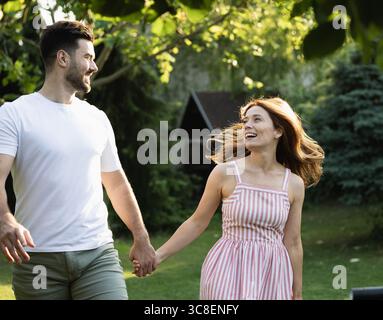 Niedliches Paar, das sich liebevoll in die Augen schaut und die Hände hält, während es an einem sonnigen Tag im Park herumläuft Stockfoto