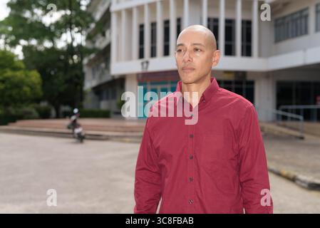 Stilvoller, kahlköpfiger italienischer Mann in burgunderfarbenem Button-Down-Hemd, der sich selbstbewusst vor dem modernen Universitätsgebäude an sonnigen Sommertagen posiert. Städtisch Stockfoto