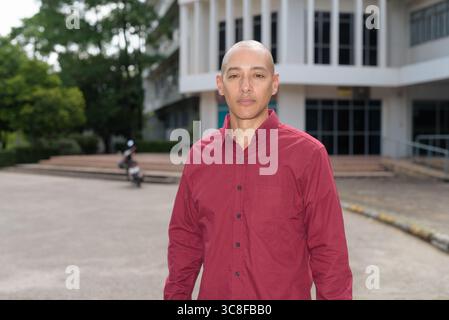 Stilvoller, kahlköpfiger italienischer Mann in burgunderfarbenem Button-Down-Hemd, der sich selbstbewusst vor dem modernen Universitätsgebäude an sonnigen Sommertagen posiert. Städtisch Stockfoto
