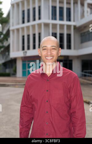 Stilvoller, kahlköpfiger italienischer Mann in burgunderfarbenem Button-Down-Hemd, der sich selbstbewusst vor dem modernen Universitätsgebäude an sonnigen Sommertagen posiert. Städtisch Stockfoto