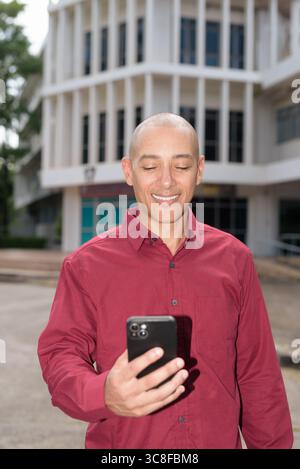 Glatzkopf italienischer Mann in burgunderrotem Hemd, der sein Smartphone überprüft, während er vor dem Universitätsgebäude bei hellem Sonnenlicht steht. Das Konzept des digitalen Kombi Stockfoto