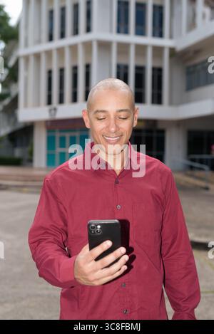 Glatzkopf italienischer Mann in burgunderrotem Hemd, der sein Smartphone überprüft, während er vor dem Universitätsgebäude bei hellem Sonnenlicht steht. Das Konzept des digitalen Kombi Stockfoto