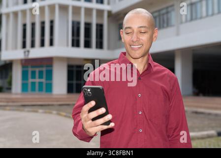 Glatzkopf italienischer Mann in burgunderrotem Hemd, der sein Smartphone überprüft, während er vor dem Universitätsgebäude bei hellem Sonnenlicht steht. Das Konzept des digitalen Kombi Stockfoto
