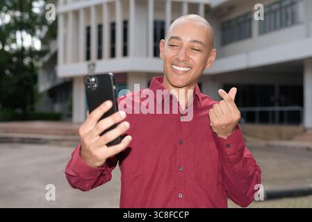 Glatzkopf italienischer Mann in burgunderrotem Hemd, der sein Smartphone überprüft, während er vor dem Universitätsgebäude bei hellem Sonnenlicht steht. Das Konzept des digitalen Kombi Stockfoto