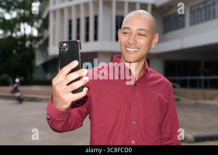 Glatzkopf italienischer Mann in burgunderrotem Hemd, der sein Smartphone überprüft, während er vor dem Universitätsgebäude bei hellem Sonnenlicht steht. Das Konzept des digitalen Kombi Stockfoto
