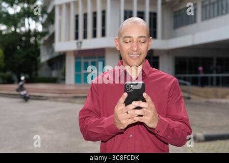 Glatzkopf italienischer Mann in burgunderrotem Hemd, der sein Smartphone überprüft, während er vor dem Universitätsgebäude bei hellem Sonnenlicht steht. Das Konzept des digitalen Kombi Stockfoto