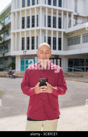 Glatzkopf italienischer Mann in burgunderrotem Hemd, der sein Smartphone überprüft, während er vor dem Universitätsgebäude bei hellem Sonnenlicht steht. Das Konzept des digitalen Kombi Stockfoto