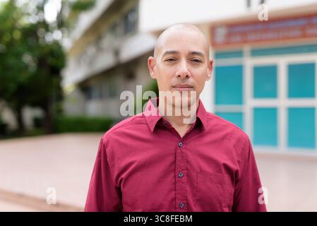 Stilvoller, kahlköpfiger italienischer Mann in burgunderfarbenem Button-Down-Hemd, der sich selbstbewusst vor dem modernen Universitätsgebäude an sonnigen Sommertagen posiert. Städtisch Stockfoto