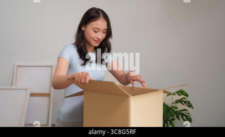 Die fröhliche junge asiatische Frau packt einen braunen Karton in ihrer modernen Wohnung aus. Sie erwartet Entdeckungen aus einer Online-Shopping-Lieferung. Stockfoto