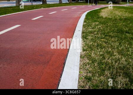 Der sich windende rote Radweg schlängelt sich durch einen Park, der von weißem Beton umgeben ist und von üppigem grünem Gras umgeben ist, lädt zum Entspannen im Freien ein Stockfoto