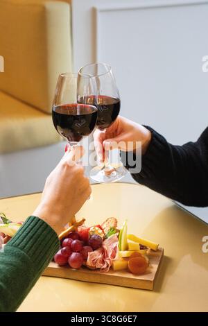 Nahaufnahme von zwei Personen, die bei einem feierlichen Toast Gläser Rotwein anstoßen, mit einer Wurstplatte mit Trauben, Käse, Wurstwaren, Stockfoto