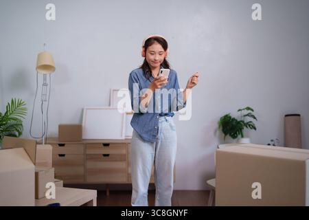Asiatische Frau, die sich während des Umzugs entspannt und Musik am Telefon genießt, neue Wohnräume und modernen Wohnstil aufbaut Stockfoto