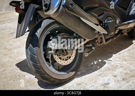 Nahaufnahme des Hinterrads eines modernen Motorrads mit Bremsscheibe, Auspuff und dickem Reifenprofil. Bei Tageslicht auf Beton geschossen. Stockfoto