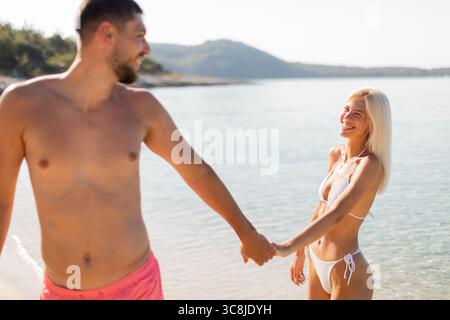 Ein Paar geht Hand in Hand an einem sonnigen Strand entlang, lächelt auf den Gesichtern. Wellen krachen sanft am Ufer, während die warme Sonne auf dem Wasser glitzert, cre Stockfoto
