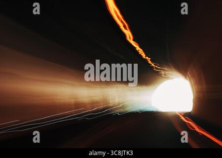 Langbelichtungsaufnahme eines Tunnels mit Lichtspuren und hellem Lichtaustritt, die eine dynamische Bewegungsunschärfe und einen dramatischen visuellen Effekt erzeugt. Selektiver Fokus. Stockfoto