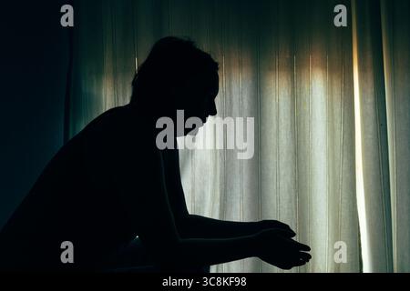 Silhouette einer Frau, die in einem dunklen Raum in der Nähe eines Fensters sitzt, Hände vor ihr gefesselt, um emotionalen Kampf oder Besinnung auszudrücken. Selektiv f Stockfoto