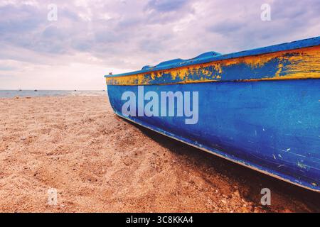 Ein verwittertes blaues Fischerboot sitzt an einem Sandstrand unter bewölktem Himmel. Selektiver Fokus. Stockfoto