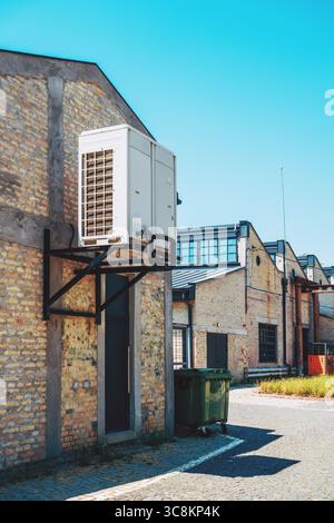 Wärmepumpeneinheit für den Außenbereich, montiert auf einem Industriegebäude aus Ziegelsteinen, mit blauem Himmel und sauberer Kopfsteinpflasterstraße, die HLK-Systeme in modernem Infrast repräsentiert Stockfoto