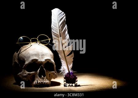Theater- oder Theaterkonzept. Alter Tintenständer mit Federfeder in der Nähe des Schädels mit alter Retro-Brille auf schwarzem Hintergrund. Dramatisches Licht. Stockfoto