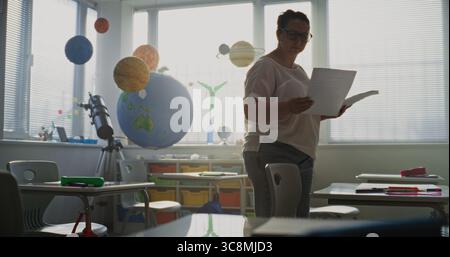 Lehrerin legt Arbeitsblätter mit Schreibübungen für die Schüler vor dem Unterricht an. Lehrer organisieren Raum für Schulprüfung im Klassenzimmer, Arbeit in der modernen Grundschule. Dolly Shot. Stockfoto