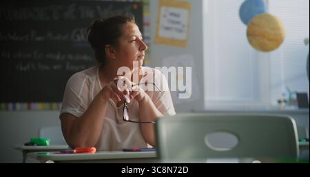 Müde Lehrerin, die allein am Schreibtisch im leeren Klassenzimmer sitzt und sich nach dem Unterricht entspannt. Frau, die Stress, Burnout und geistige Erschöpfung im Bildungsumfeld empfindet, Arbeit in der Grundschule. Stockfoto